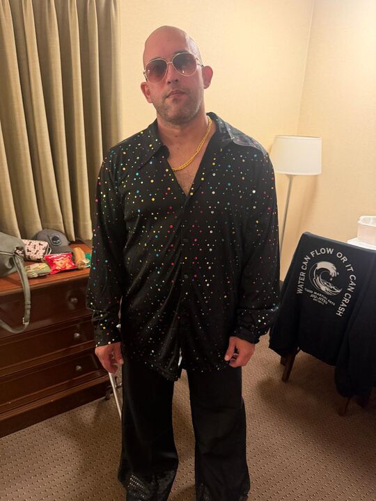 Commander standing in a hotel room in a full black sequinned suit wearing sunglasses, before the wig was fitted