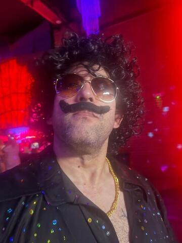 Commander in a curly black wig, large fake moustache and tinted aviators under red club lighting