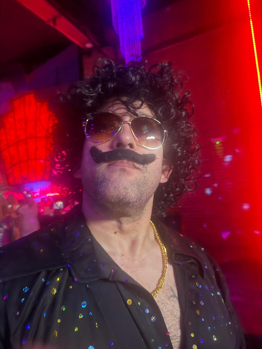 Commander in a curly black wig, oversized fake moustache and tinted aviator sunglasses, wearing an open sequinned shirt under red club lighting