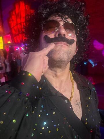 Commander pointing at his large fake moustache under red and blue club lighting