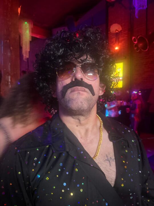 Commander facing the camera in a curly wig and large fake moustache under red club lighting