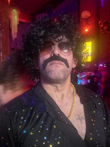 Commander facing the camera in a curly wig and fake moustache under red club lighting