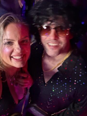 Commander in a wig and tinted glasses smiling with Night Shade in a club