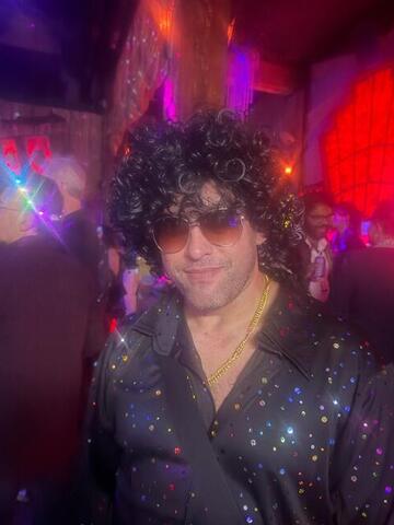 Commander standing in a curly wig and sequinned shirt on a busy club floor