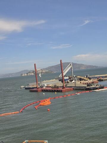 An orange floating boom and a work barge in the bay seen from above