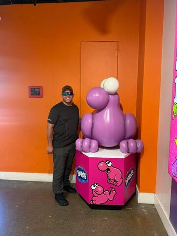 Commander standing beside a large pink candy mascot statue in a bright orange room