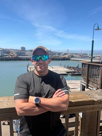 Commander standing with arms crossed at a waterfront railing wearing mirrored sunglasses