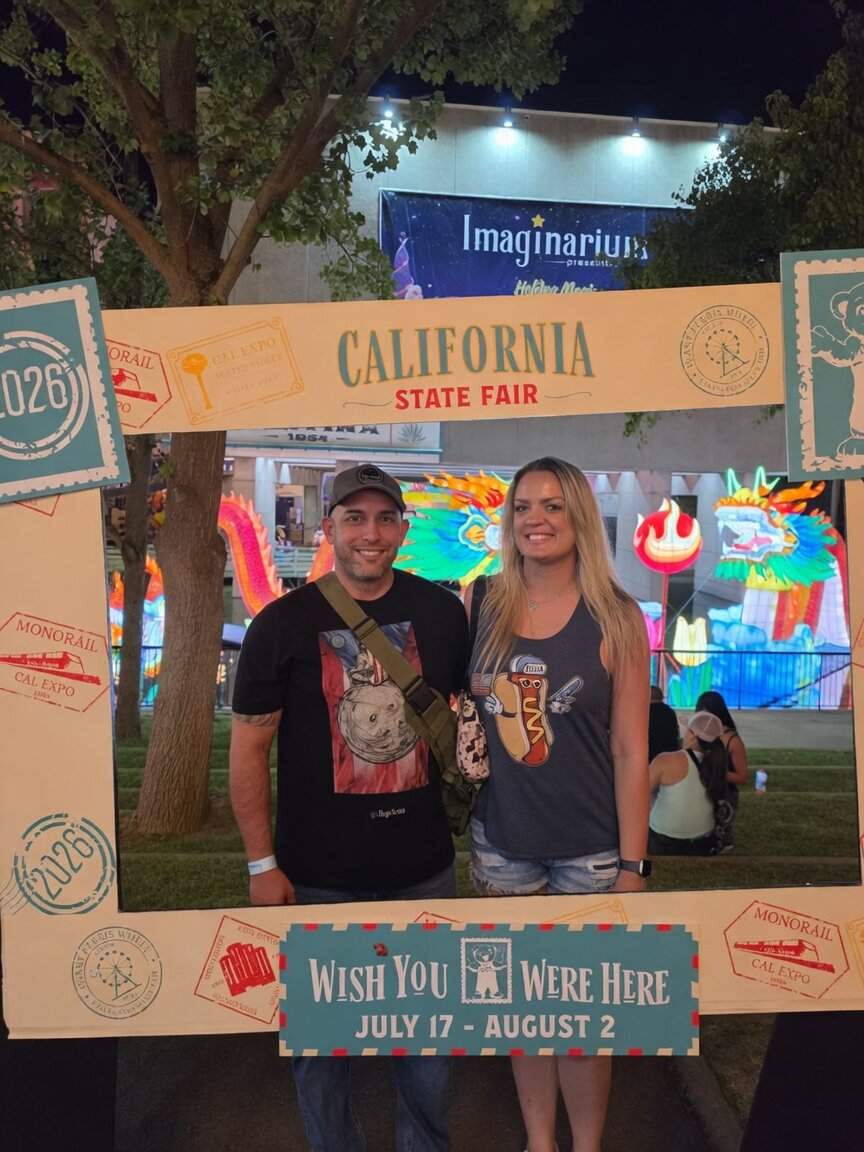 Commander and Night Shade standing together inside the California State Fair Wish You Were Here postcard frame at night, neon dragon lanterns behind them